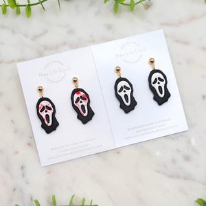 Könnte beinhalten: Zwei Paar handgefertigte Polymer Clay Ohrringe mit einem schwarz-weißen Scream-Masken-Design. Die Ohrringe haben goldfarbene Stifte und werden auf weißen Karten mit dem Logo und Text von Happy Little Clay Co. präsentiert.