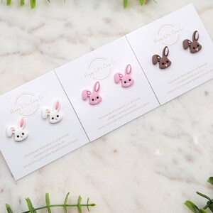 May include: Three cards with pairs of polymer clay bunny earrings. The bunny earrings are in white, pink, and brown. The cards have the text "Happy Little Clay Co" and information about the handmade jewelry.
