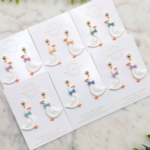 Puede incluir: Pendientes de arcilla polimérica hechos a mano con diseños de gansos blancos con lazos de colores. Cada pendiente tiene un perno dorado y patas naranjas. Las tarjetas dicen "Happy Little Clay Co."