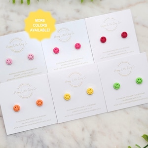May include: A selection of smiley face stud earrings in various colours, including pink, red, orange, yellow, and green, displayed on white cards. Each card features the text "Happy Little Clay Co" and "More Colors Available!"