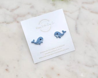 May include: A pair of light blue whale-shaped polymer clay earrings on a white card. The card has the text "Happy Little Clay Co" and other text. The earrings are on a marble surface with green leaves.