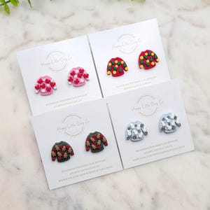 Könnte beinhalten: Vier Paar handgefertigte Ohrringe aus Polymer Clay mit verschiedenen Weihnachtspullover-Designs. Die Pullover sind rot, grün, blau und pink mit verschiedenen Mustern, darunter Schneeflocken, Rentiere und Weihnachtsbäume. Die Ohrringe werden auf weißen Karten mit dem Text "Happy Little Clay Co." präsentiert.