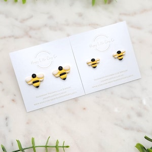 May include: Two pairs of handmade polymer clay bee earrings. The bees are yellow with black stripes and white wings. The earrings are on white cards with the text "Happy Little Clay Co" and website information.