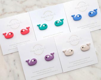 May include: A collection of handmade polymer clay whale stud earrings in various colors, including red, teal, blue, purple, and beige. Each whale features a small white flower detail. The earrings are displayed on white cards.