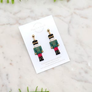 Può includere: Un paio di orecchini in argilla polimerica a forma di soldatini schiaccianoci giocattolo. Gli orecchini presentano giacche verdi, cappelli neri e stivali rossi. Accenti dorati e perni per le orecchie completano il design. Gli orecchini sono esposti su una carta bianca.