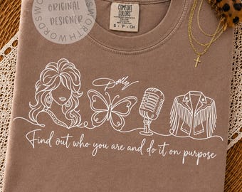 Può includere: Una t-shirt marrone con una grafica a linee bianche raffigurante una donna con capelli voluminosi, una farfalla, un microfono vintage e una giacca con frange. Il testo recita "Dolly" e "Find out who you are and do it on purpose". Una molletta per capelli leopardata poggia sul colletto.