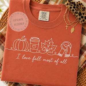 May include: A rust-colored t-shirt featuring a white line art graphic of a pumpkin, a coffee cup with a heart, a maple leaf, and a scarf. Below the graphic, the text reads, I love fall most of all. The shirt has a Comfort Colors brand label.