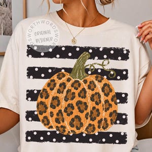 May include: A cream-colored t-shirt featuring a central graphic of a pumpkin with an orange and black leopard print pattern. The pumpkin is set against a background of horizontal black stripes with white polka dots and a circular logo text.
