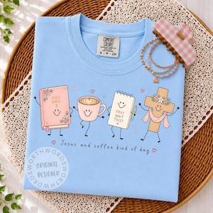 May include: A light blue t-shirt featuring a graphic of a smiling Holy Bible, a coffee cup, a notepad with the text PRAY WAIT TRUST, and a cross with a floral crown. The text Jesus and coffee kind of day is printed below the characters.
