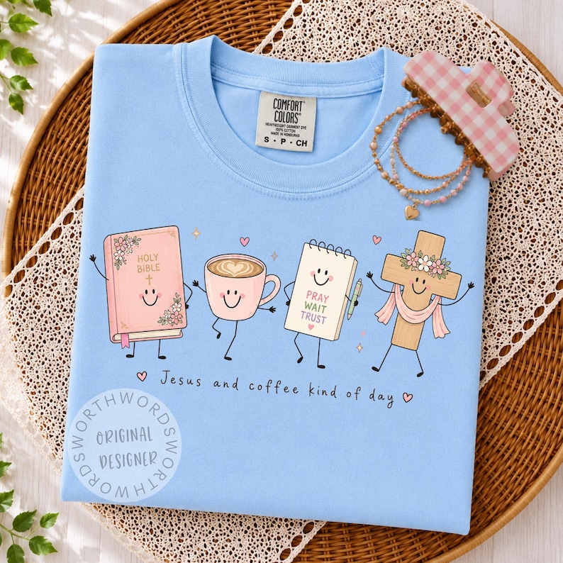 May include: A light blue t-shirt featuring a graphic of a smiling Holy Bible, a coffee cup, a notepad with the text PRAY WAIT TRUST, and a cross with a floral crown. The text Jesus and coffee kind of day is printed below the characters.