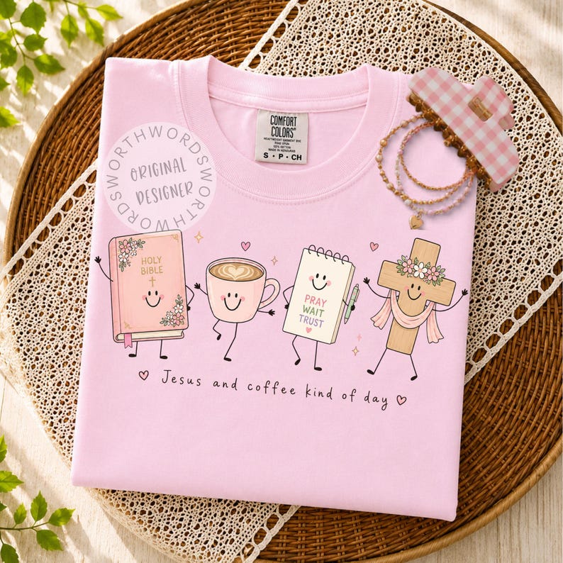 May include: A light pink t-shirt featuring a graphic of a smiling Holy Bible, a coffee cup, a notepad with the text PRAY WAIT TRUST, and a cross. The text Jesus and coffee kind of day is printed below the characters. A pink gingham hair clip and beaded bracelets are placed on the shirt.