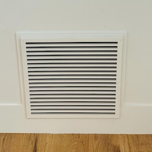 Op de afbeelding: Een witte rechthoekige ventilatieopening met horizontale zwarte lamellen is in een witte muur geplaatst. De ventilatieopening bevindt zich boven een houten vloer. De muur heeft een witte plint. Een raam is linksboven zichtbaar.