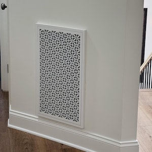 May include: A rectangular white wall vent cover featuring a repeating geometric pattern of small black squares. The vent is mounted flush against a smooth white wall, positioned above the baseboard in a home interior setting with wooden flooring.