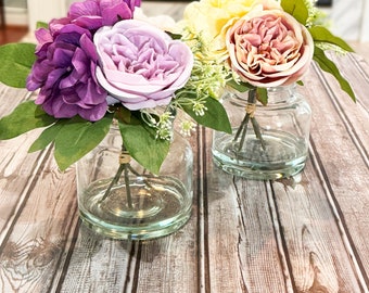 May include: Two glass vases with floral arrangements. The vase on the left has purple and pink flowers, while the vase on the right has yellow and pink flowers. The vases are sitting on a wooden surface.