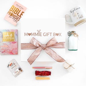 May include: A white gift box with a rose gold ribbon, surrounded by various items. Includes a mug with text, a journal, a book titled "Bible Promises for Moms", a bottle, a pen, and a bundle of sage.