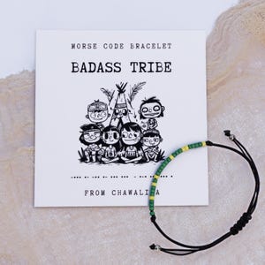 May include: A black cord bracelet with green and yellow beads, next to a white card. The card features a black and white illustration of cartoon characters and the text "MORSE CODE BRACELET BADASS TRIBE FROM CHAWALI A".