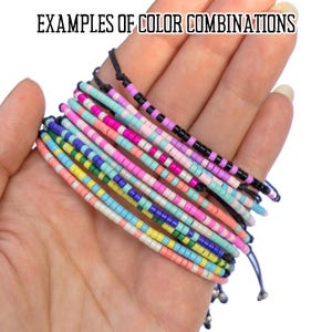 May include: A close-up of a hand holding a stack of colorful beaded bracelets. The bracelets are made with small, round beads in various colors, including pink, blue, green, yellow, and white. The bracelets are tied together with black string.