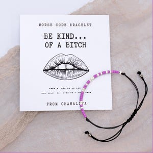 May include: A purple and white beaded bracelet with a black cord and silver beads. The bracelet is on a white card with the text "Morse Code Bracelet", "Be Kind... Of A Bitch", and "From Chawalita".