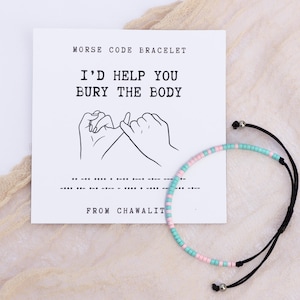 May include: A pink and blue beaded bracelet with a black cord. The bracelet is on a white card with the text "Morse Code Bracelet" and "I'd help you bury the body" in black text. The card also includes a drawing of two hands making a pinky promise and the morse code translation of the text.