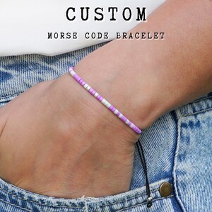 May include: A purple and white beaded bracelet with a black cord. The bracelet is on a person's wrist and the cord is hanging down. The text "CUSTOM MORSE CODE BRACELET" is at the top of the image.
