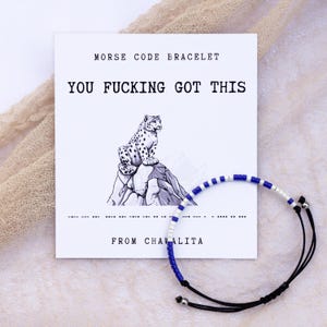 May include: A white card with the text "Morse Code Bracelet" and "You Fucking Got This" in black text. A black and white illustration of a snow leopard on a mountain is below the text. A blue and white beaded bracelet with black string is on the right side of the card. The text "From Chavalita" is below the bracelet.