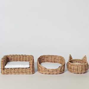 May include: Three small pet beds made of light brown woven wicker. The first is rectangular with a white cushion, the second is oval with a white cushion, and the third is round with two small pointed ears on the rim and a white cushion.