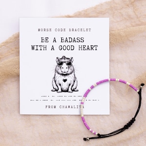 May include: A white card with the text "Morse Code Bracelet" and "Be a badass with a good heart". A cartoon image of a rodent wearing sunglasses and a heart on its chest is in the center of the card. A purple and white beaded bracelet with a black cord is on the right side of the card. The text "From Chawalita" is below the morse code.