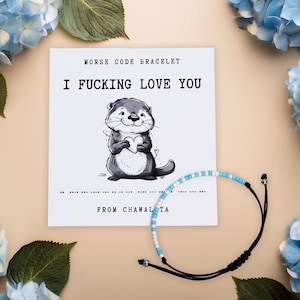 May include: A white card with the text "Morse Code Bracelet" and "I FUCKING LOVE YOU" in black text. A cartoon otter holding a heart is pictured below the text. A blue and white beaded bracelet with black string ties is shown next to the card.