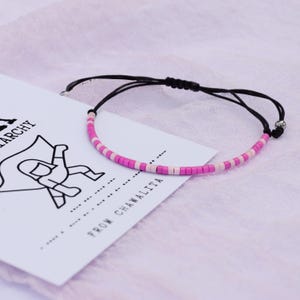 May include: A pink and white beaded bracelet with a black cord and silver bead accents. The bracelet is on a white card with the text "From Chawalita" printed on it.
