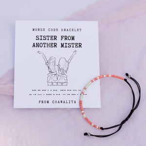 May include: A pink and white beaded bracelet with a black cord. The bracelet is on a white card with the text "Morse Code Bracelet Sister From Another Mister From Chawalita".