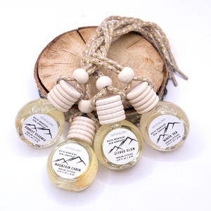 May include: Five small, round hanging diffusers with wooden tops and labels. The labels read "Forest Essence", "Mountain Cabin", "Citrus Glow", and "White Tea". The diffusers are suspended from a braided rope and rest on a wooden slice.