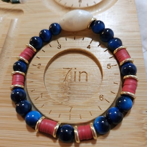 Op de afbeelding: Een kralenarmband met afwisselend donkerblauwe ronde kralen, rode schijfvormige kralen en een enkele witte kaurischelp. De armband ligt op een houten meetinstrument dat een omtrek van 17,78 cm aangeeft.
