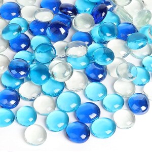 May include: A collection of smooth, rounded glass gems in various shades of blue, light blue, and clear white. These circular, flat-bottomed decorative stones are scattered across the frame, displaying translucent qualities and reflective surfaces.