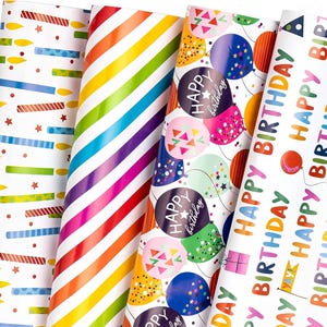 Peut inclure: Cinq rouleaux de papier cadeau aux motifs variés. On y trouve des ballons d'anniversaire, des bougies et des rayures arc-en-ciel. Le papier porte l'inscription "Happy Birthday" en plusieurs couleurs.