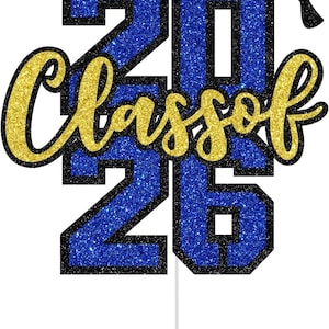 Puede incluir: Un adorno para pastel de graduación con el texto "Class of 2026". Los números y el texto son de purpurina dorada con contorno negro, sobre un fondo de purpurina azul. Una birrete negro en la parte superior.