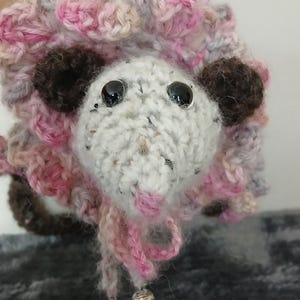 Könnte beinhalten: Ein gehäkeltes Stofftier mit einem Farbschema aus Rosa, Weiß und Braun. Das Tier hat große schwarze Augen und eine rosa Nase. Das Tier hat eine flauschige Textur und trägt eine rosa-weiße Häkelmütze.