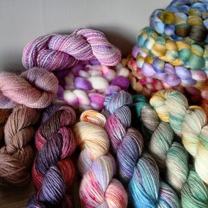 Könnte beinhalten: Eine Sammlung bunter Garnknäuel in verschiedenen Schattierungen von Lila, Rosa, Braun, Blau und Grün. Das Garn ist kreisförmig angeordnet, wobei sich einige Knäuel überlappen.