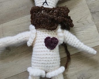 May include: White crocheted stuffed animal with a black beret, brown scarf, and a purple heart on its chest. The animal has black eyes and a sad expression.