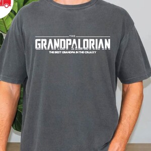 May include: A dark gray t-shirt with the text "THE GRANDPALORIAN THE BEST GRANDPA IN THE GALAXY" printed in white.