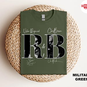 May include: Military green t-shirt featuring the text "R&B" with images of Usher Raymond and Chris Brown. The shirt includes the artists' signatures. The shirt is displayed on a woven surface.