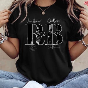 May include: Black t-shirt featuring the text "R&B" with images of Usher Raymond and Chris Brown. The design includes their names and signatures in white. The shirt is worn by a person.