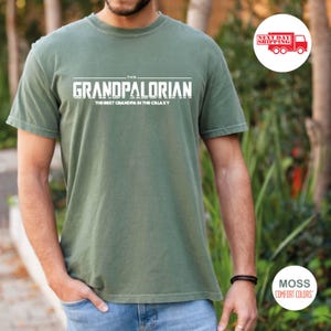May include: A green t-shirt with white text that reads "The Grandpalorian, The Best Grandpa in the Galaxy".
