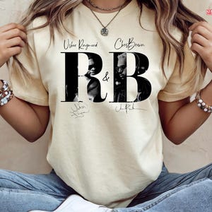 May include: Cream-colored t-shirt featuring the text "Usher Raymond & Chris Brown R&B" in a bold, black font. The design includes black and white images of the artists and their signatures. The shirt is a casual wear item.