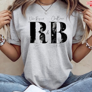 May include: A light gray t-shirt featuring a black and white graphic of Usher Raymond and Chris Brown with the letters "RB" in a bold font. The shirt includes the artists' signatures.