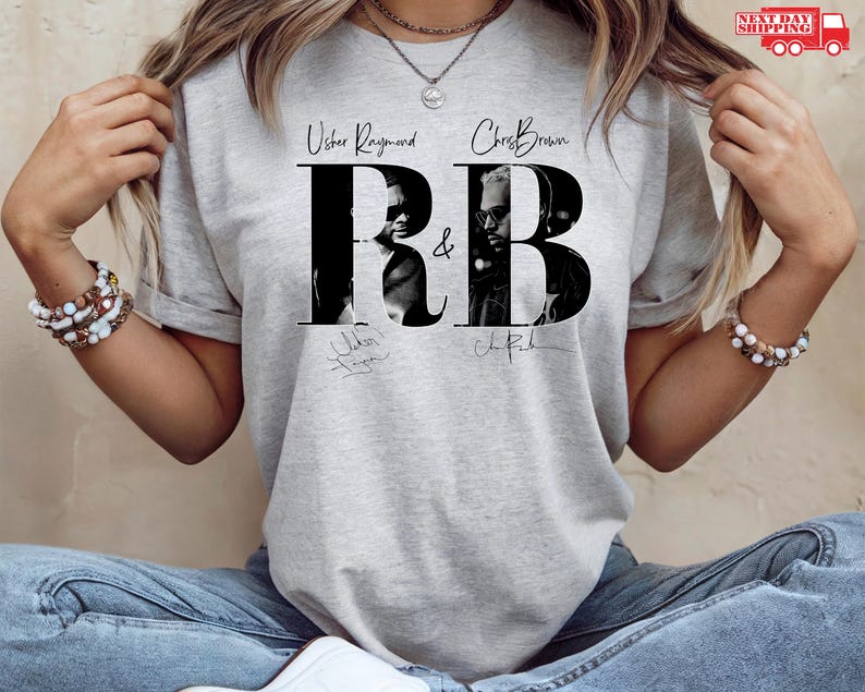 May include: A light gray t-shirt featuring a black and white graphic of Usher Raymond and Chris Brown with the letters "RB" in a bold font. The shirt includes the artists' signatures.