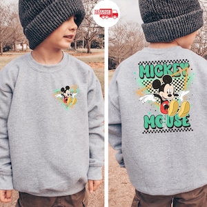 Puede incluir: Una sudadera gris con Mickey Mouse. La parte delantera tiene un peque&ntilde;o gr&aacute;fico de Mickey, mientras que la parte trasera muestra un dise&ntilde;o m&aacute;s grande con "Mickey Mouse" en letras verdes. El dise&ntilde;o incluye un patr&oacute;n de cuadros y acentos coloridos.