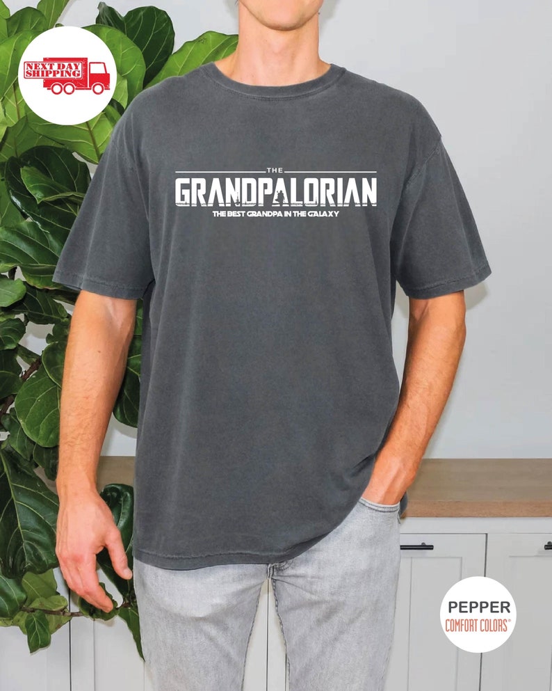 May include: A dark gray t-shirt with the text "THE GRANDPALORIAN THE BEST GRANDPA IN THE GALAXY" printed in white.