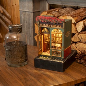 May include: A miniature model of "Taylor's Library" with a detailed interior, lit from within. The facade is green with red brick accents and a floral awning. A glass jar with a metal cage sits to the left.