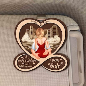 May include: A heart-shaped car visor clip with a couple embracing in front of a city skyline. The clip has the names "Linda" and "Lucas" and the phrases "I Fucking Love You" and "Drive Safe."