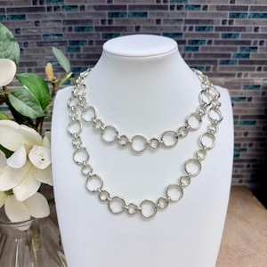 Peut inclure: Un collier argenté à double rangée. Le collier présente une série de maillons circulaires interconnectés, créant un look audacieux et élégant. Le bijou est présenté sur un mannequin blanc, mettant en valeur son design et son style. L'arrière-plan présente un motif de carreaux de mosaïque.
